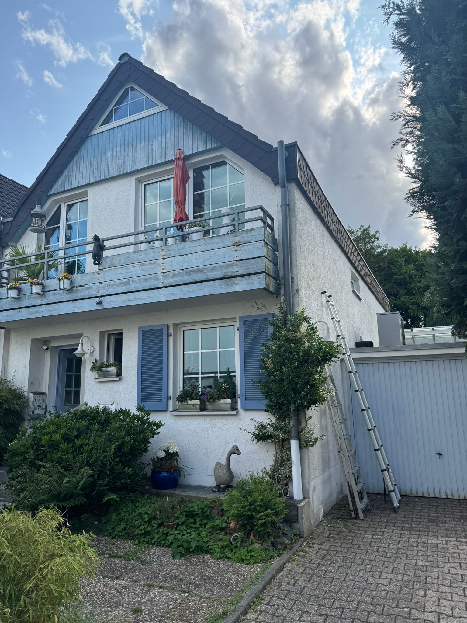 Weißes Einfamilienhaus mit Dachgaube, Balkon, Garten und Garage, Leiter an der Seitenwand angelehnt.